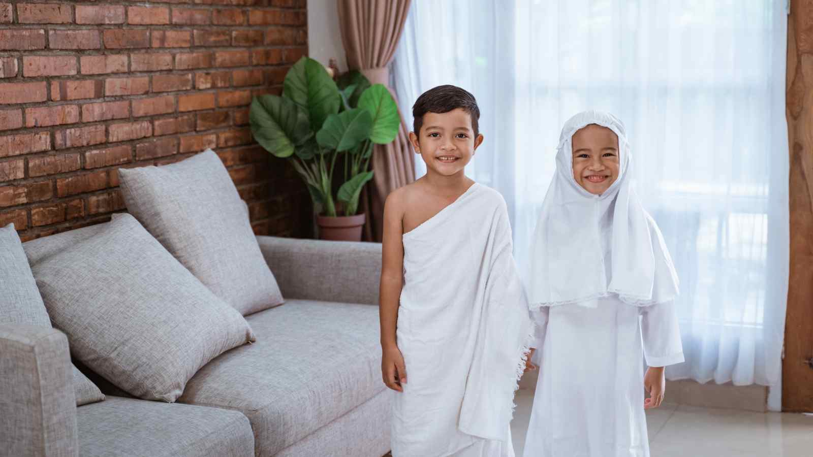 Two young children stand indoors; the boy wears a white cloth and the girl wears a white dress with a headscarf. They are smiling and holding hands in a living room.