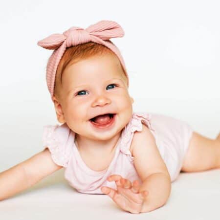 A smiling baby girl, a perfect inspiration for girl names that start with R, wears a pink headband and light outfit as she lies on her stomach against a plain white background.