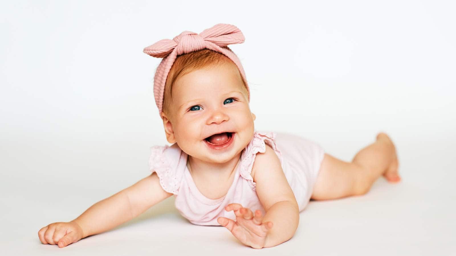 A smiling baby girl, a perfect inspiration for girl names that start with R, wears a pink headband and light outfit as she lies on her stomach against a plain white background.