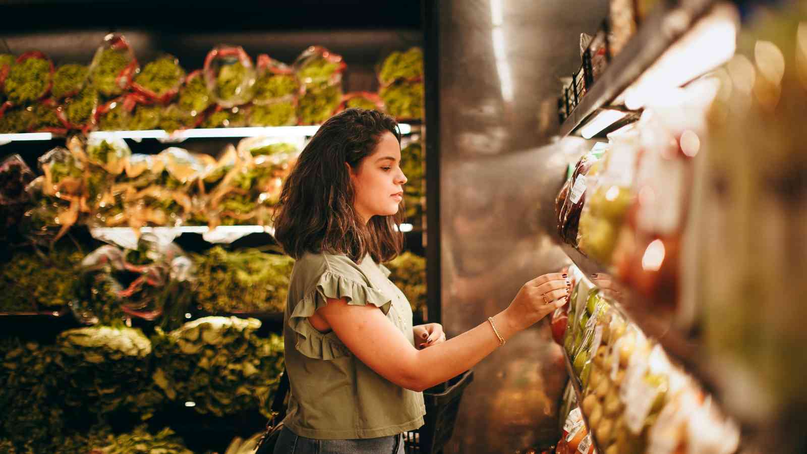 A woman stands in a grocery store produce section, selecting items from a refrigerated shelf with leafy greens visible in the background.