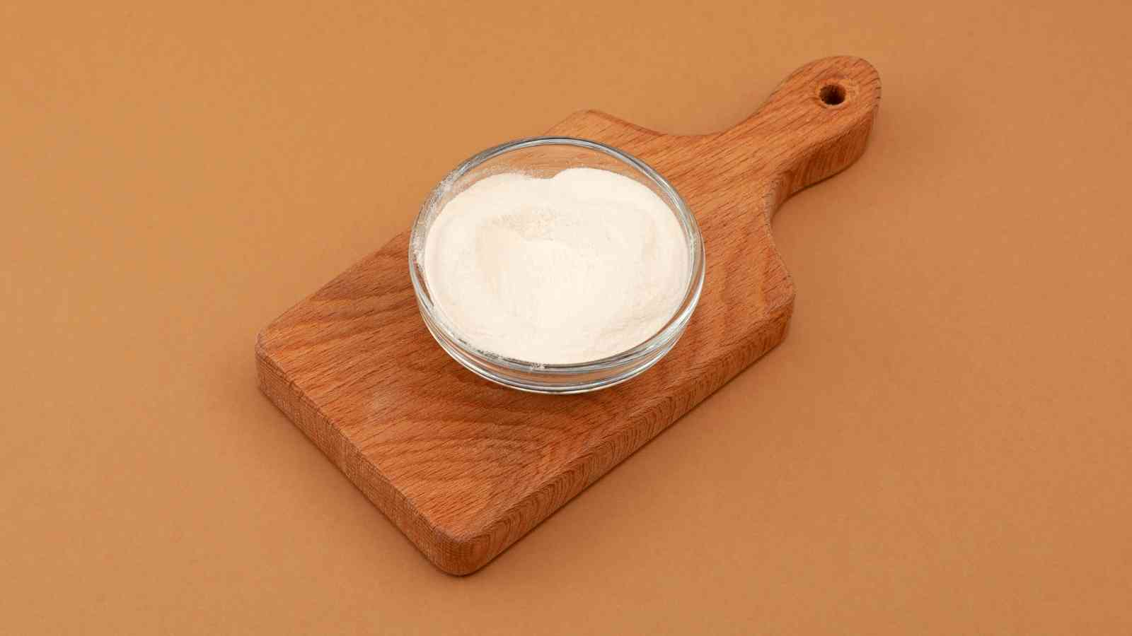A glass bowl filled with white powder sits on a small wooden cutting board against a brown background.