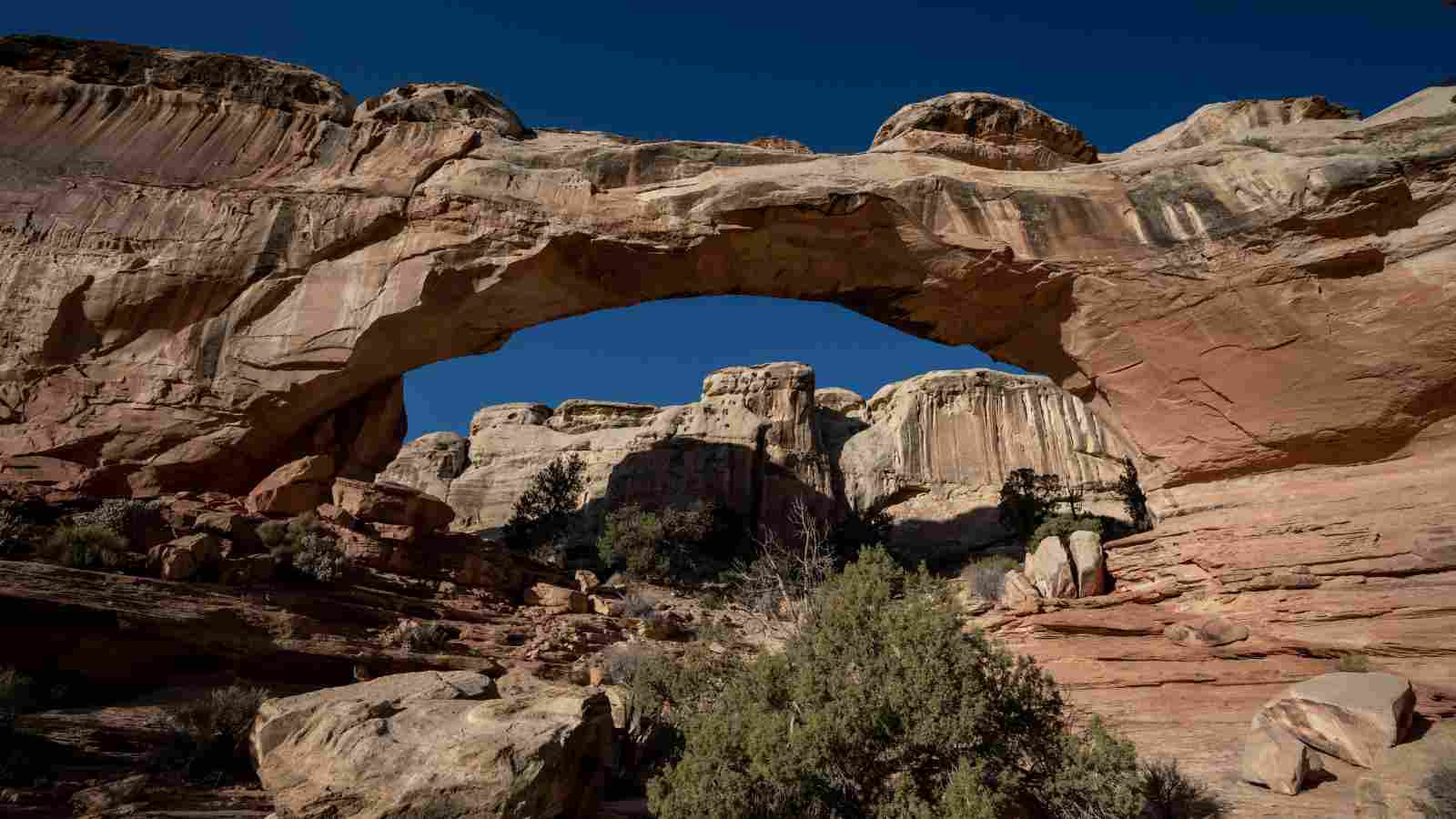 Natural rock arch formation with rugged, layered cliffs and sparse vegetation under a clear blue sky.