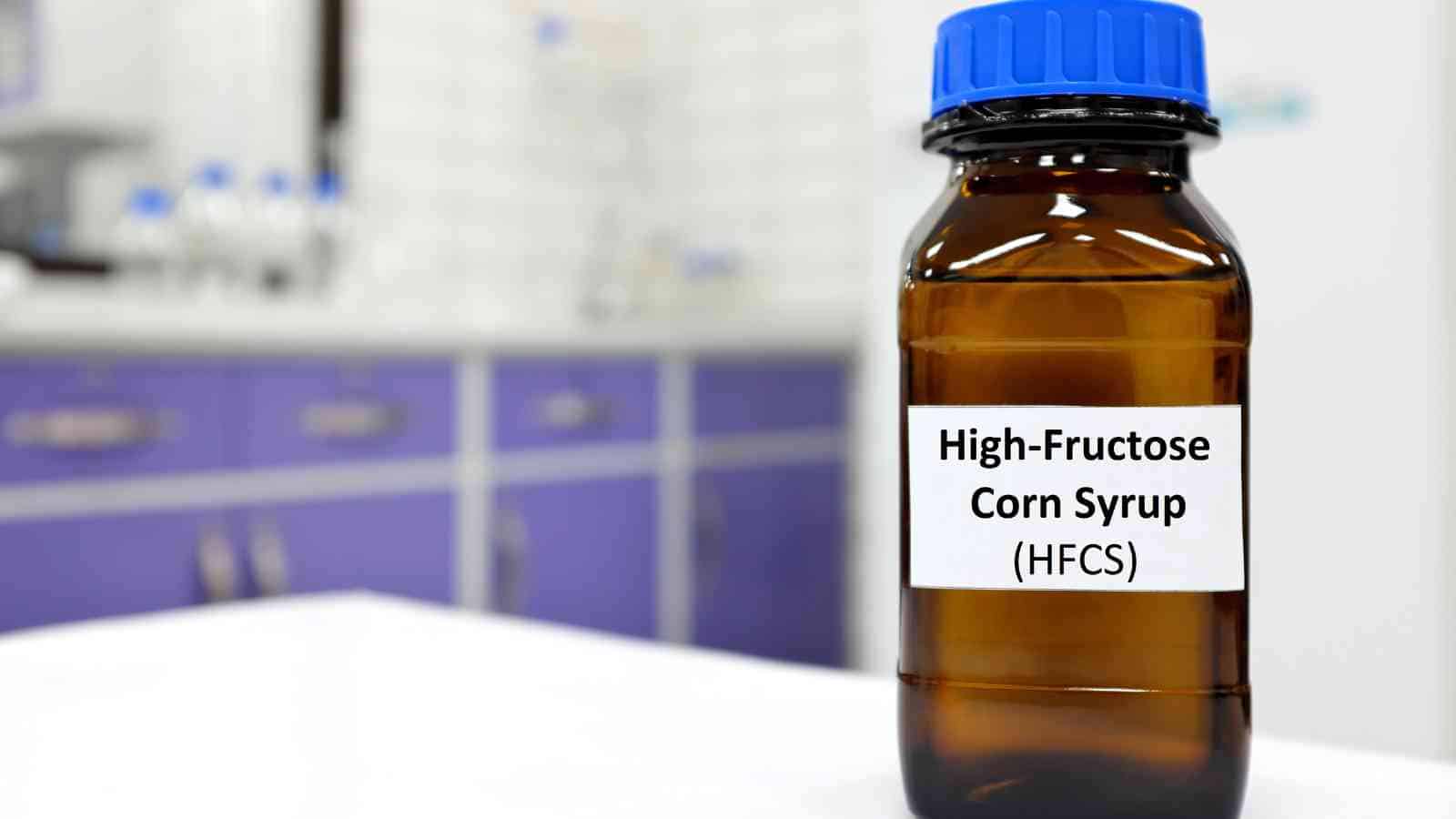 A brown lab bottle with a blue cap labeled "High-Fructose Corn Syrup (HFCS)" sits on a white surface in a laboratory setting.