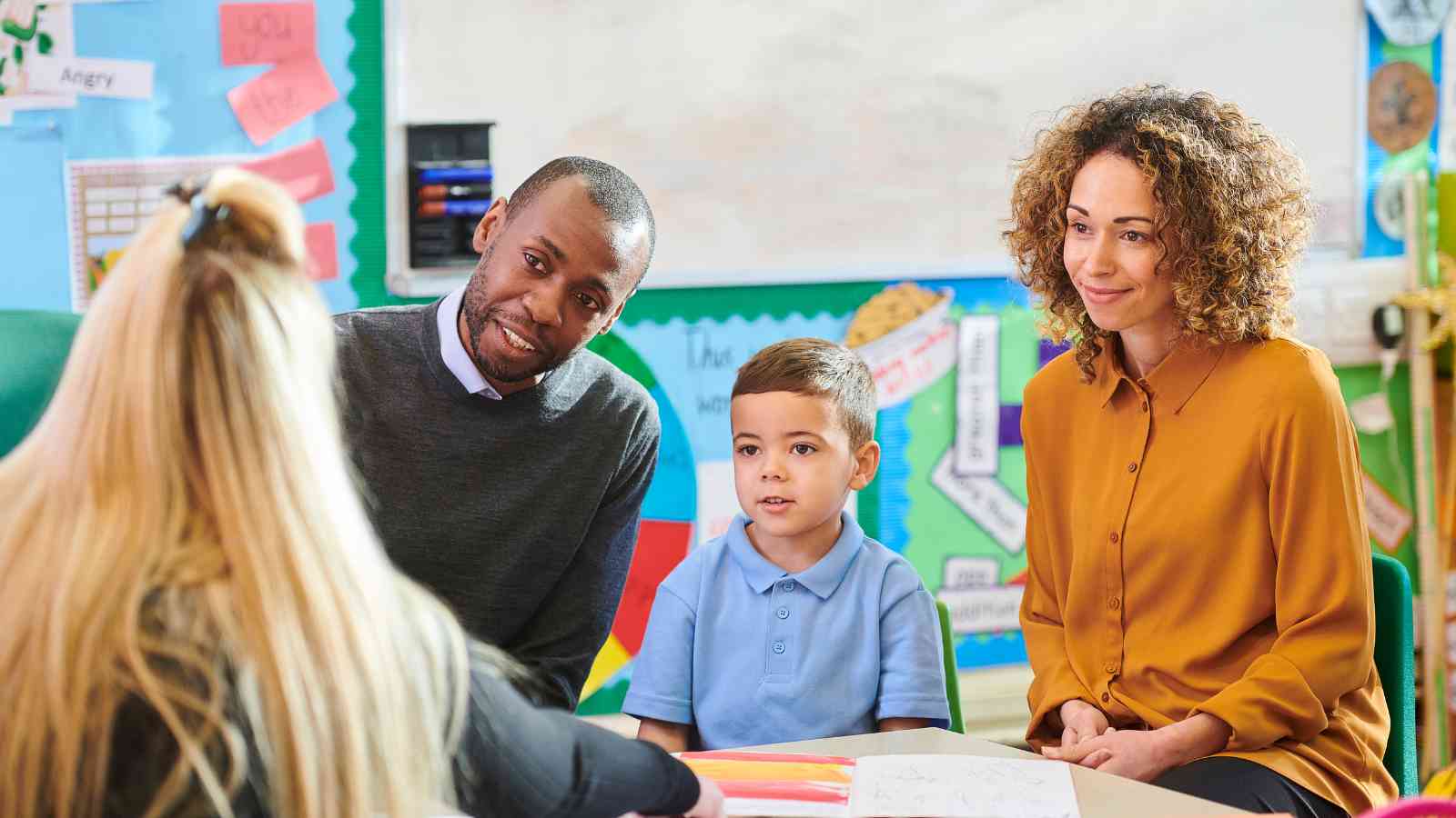 A young boy sits between two adults during a meeting with a teacher in a colorful classroom setting.