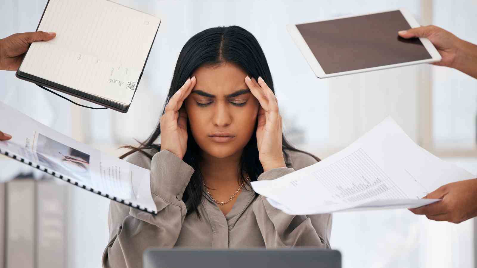 A woman sits at a desk with her eyes closed and hands on her temples, surrounded by people holding papers, a notebook, and a tablet.
