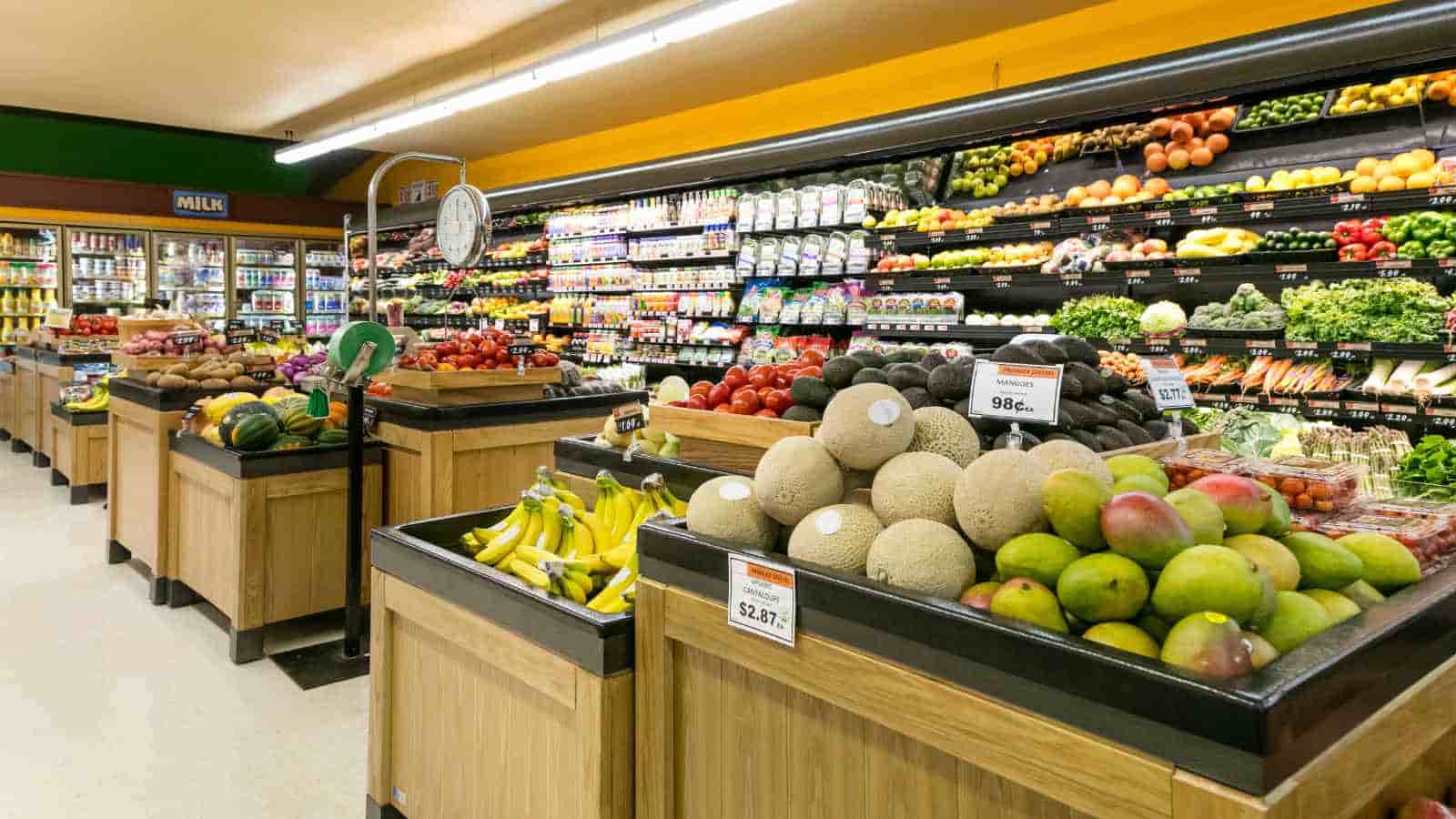 A grocery store produce section with wooden bins displaying bananas, melons, mangoes, tomatoes, and other fruits, with shelves of dairy and beverages in the background.