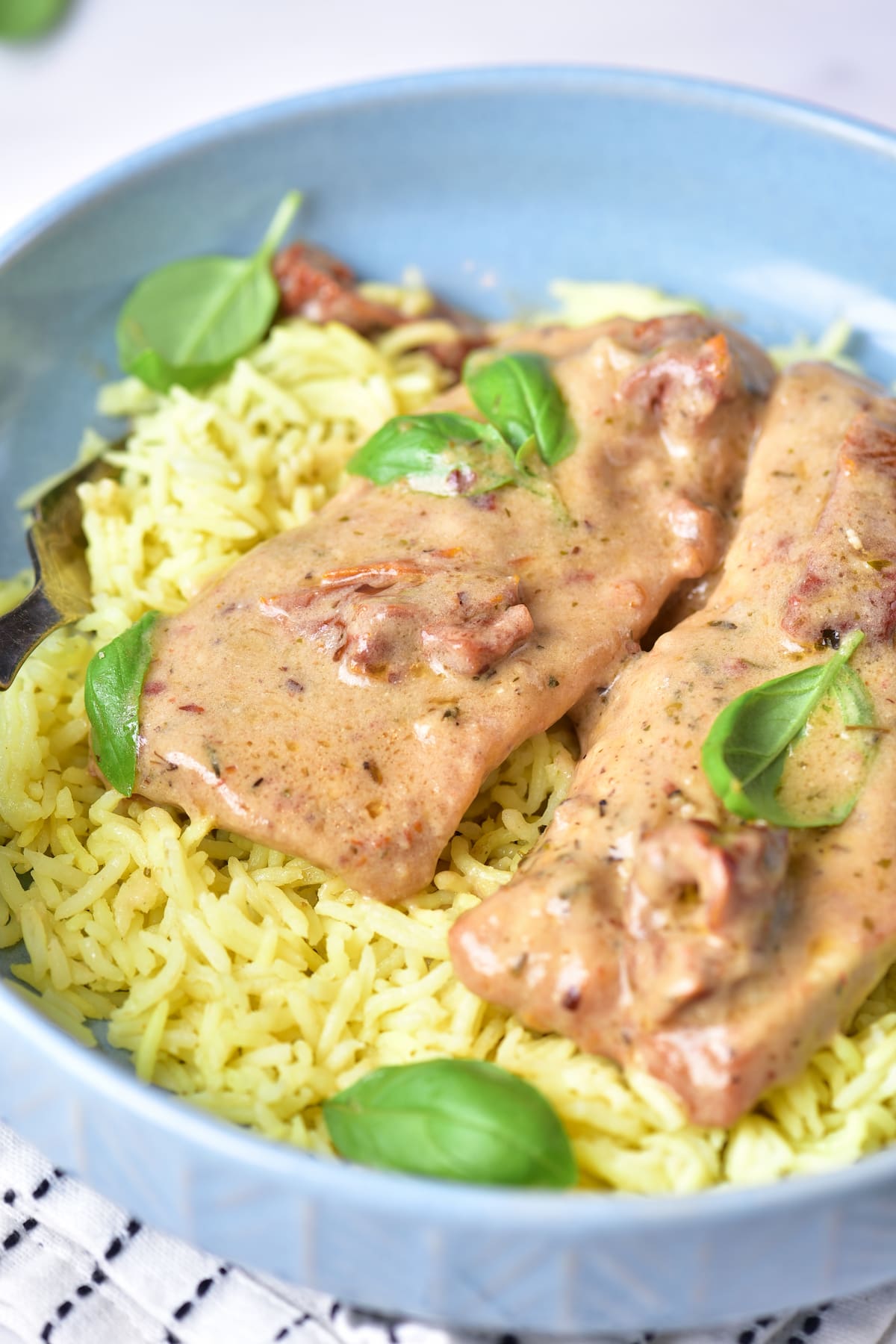 A blue bowl filled with yellow rice topped with two pieces of marry me chicken in a creamy sauce, garnished with fresh basil leaves.