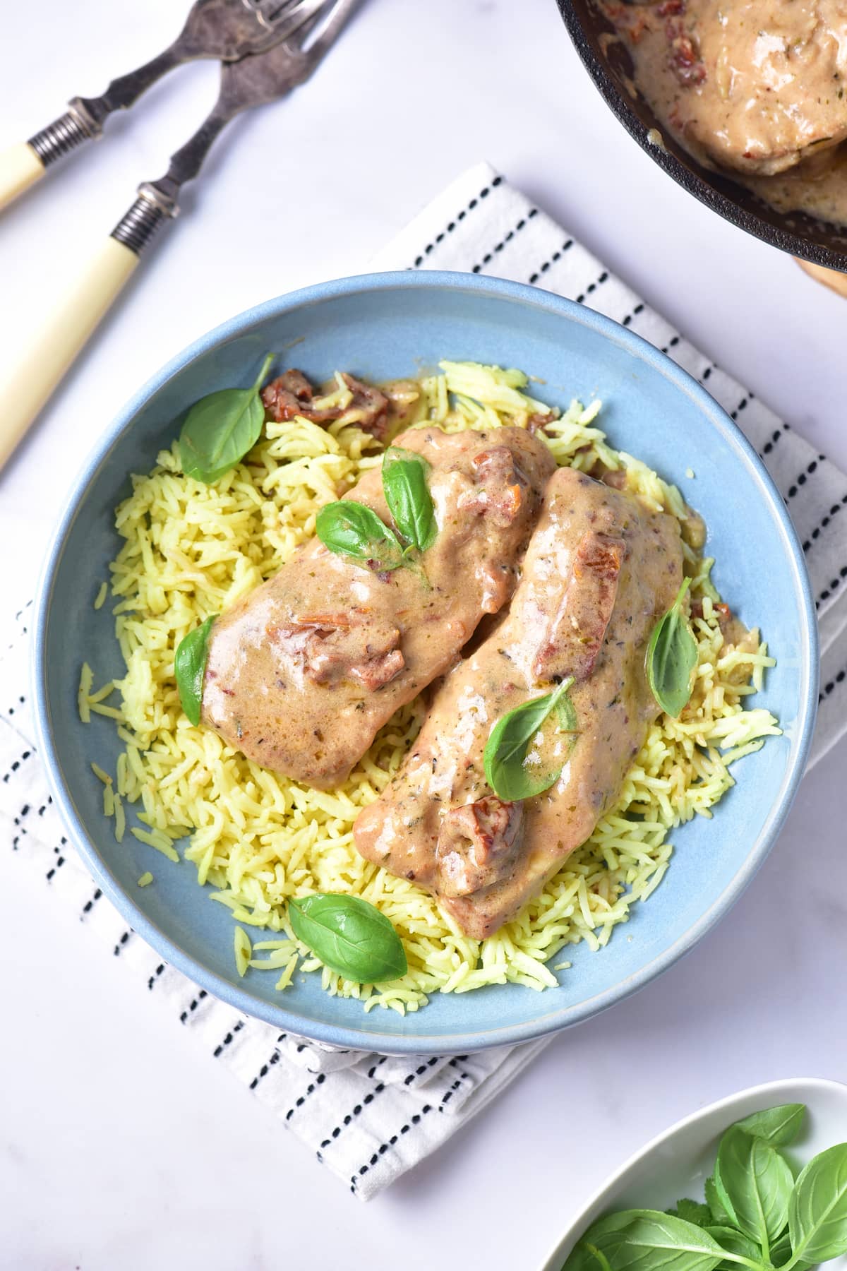 A blue bowl filled with yellow rice, topped with two pieces of creamy marry me chicken and garnished with fresh basil leaves.