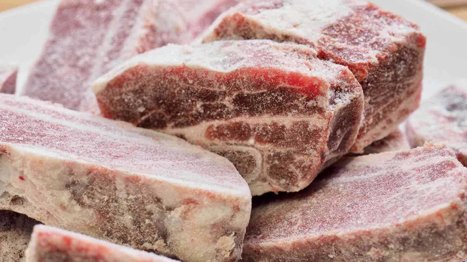 Several raw, frozen meat steaks stacked on a plate, showing visible frost and ice crystals on their surfaces.