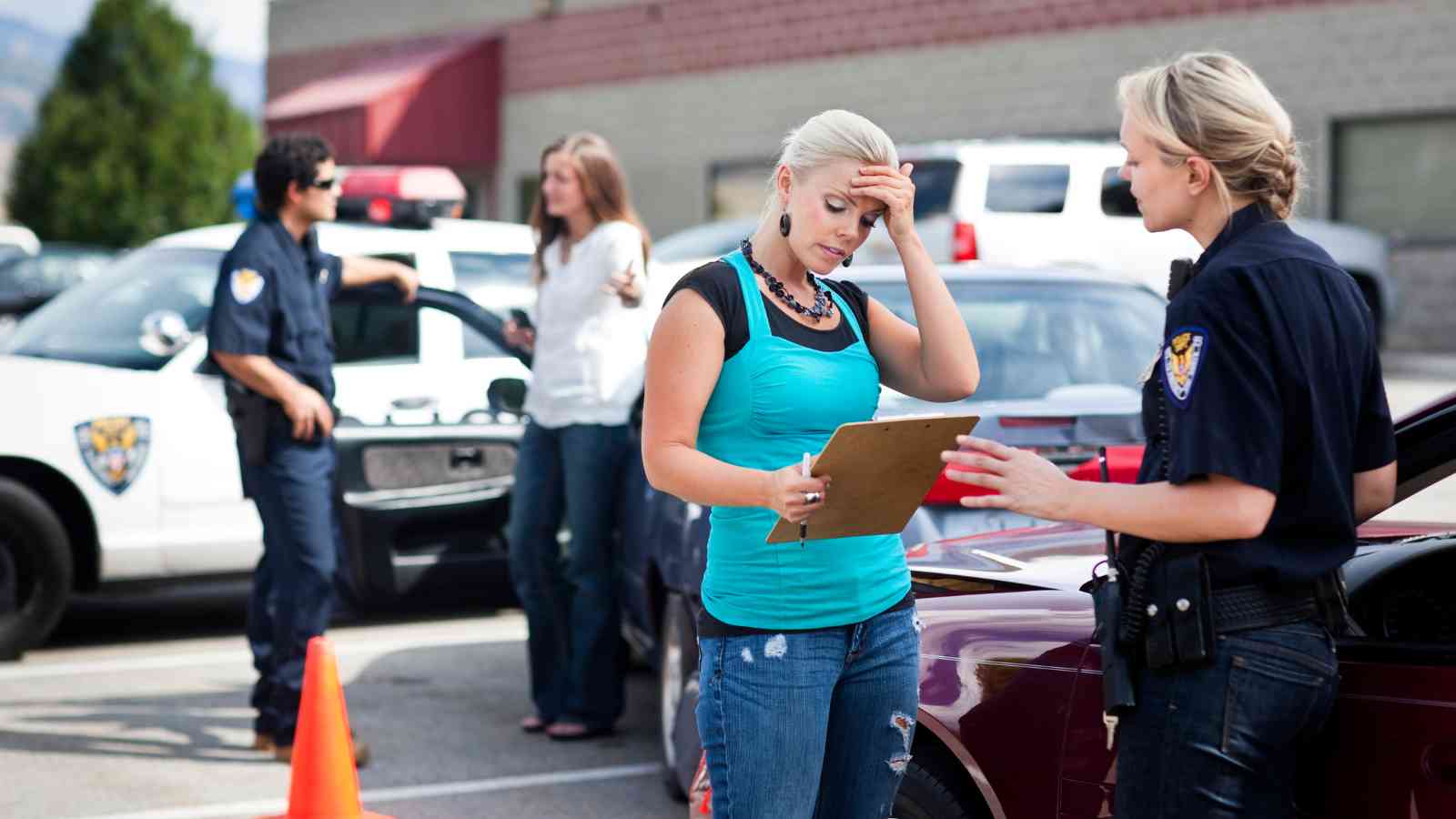 Two police officers and two women are at the scene of a car incident in a parking lot. One officer speaks to a woman holding her head while filling out paperwork.