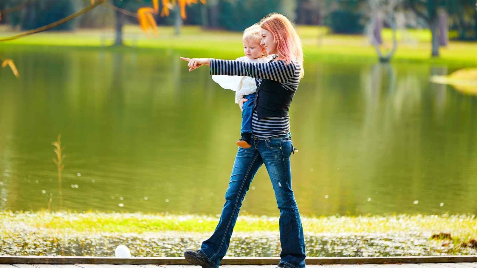 A woman holding a child points towards something across a lake in a park setting with green grass and trees in the background.