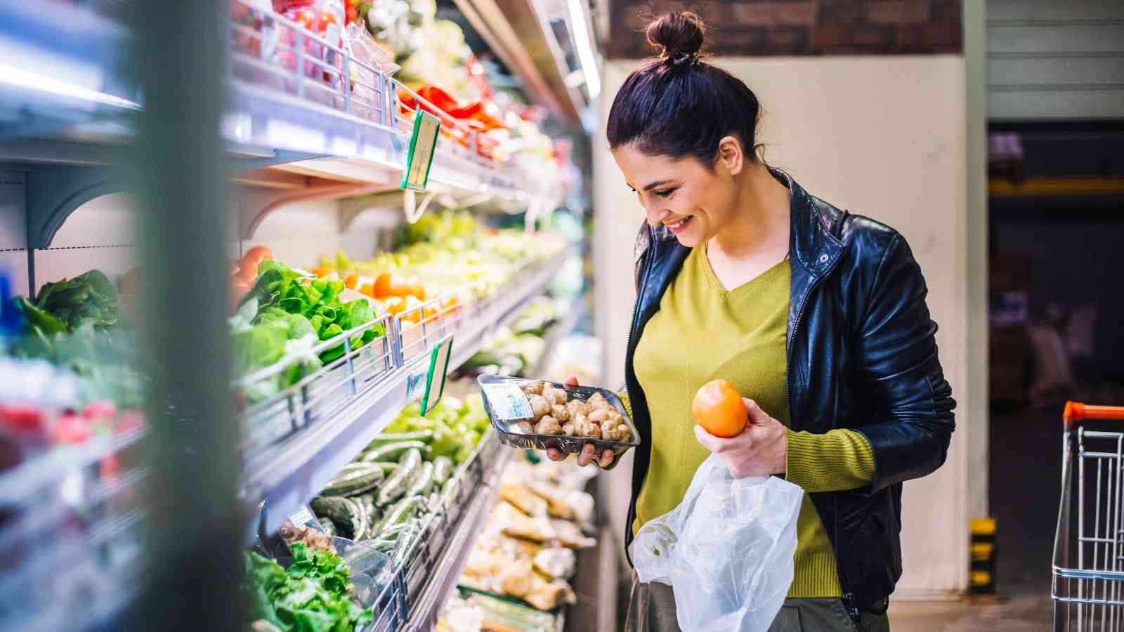 Woman shopping for fresh produce in a grocery store, holding a packaged item and an orange while looking at vegetables on the shelf.