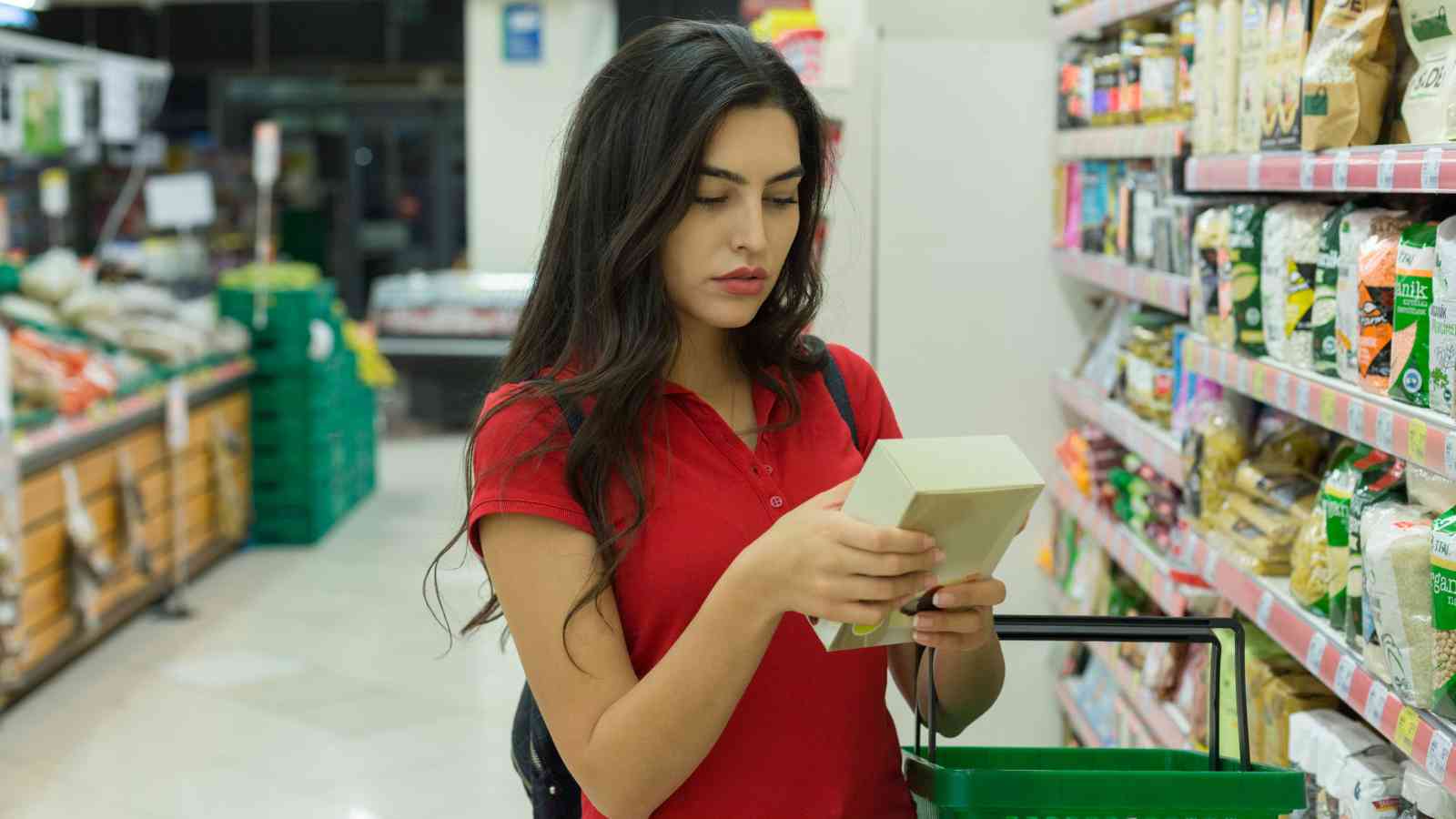 A woman in a red shirt holds a product and reads the packaging while shopping in a grocery store aisle with shelves stocked with various items.