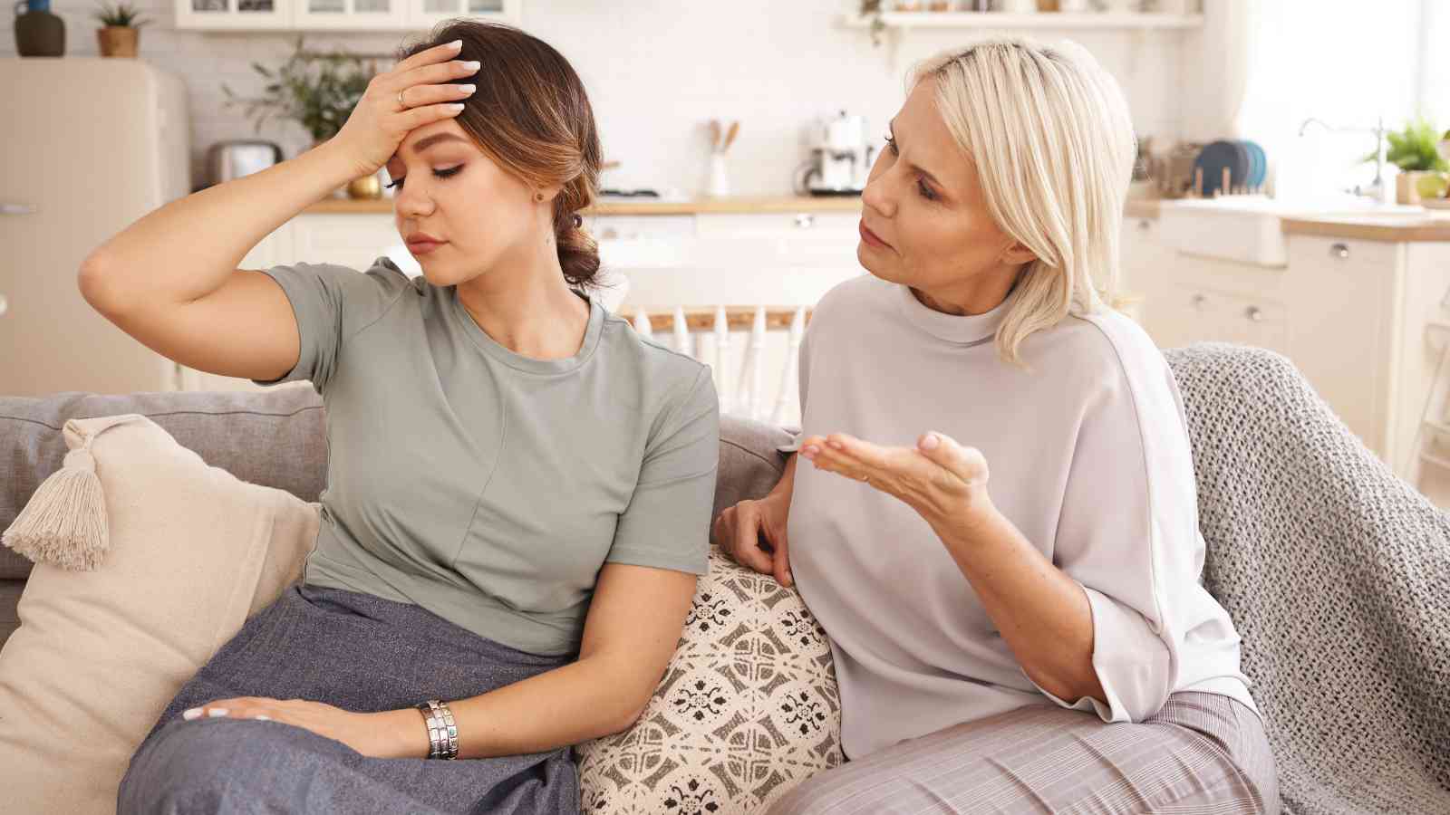Two women sit on a couch; one looks frustrated with her hand on her forehead, while the other gestures as if explaining or arguing.