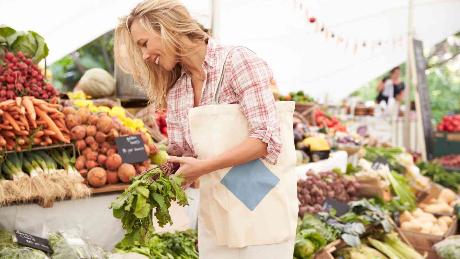 Woman holding fresh beets and shopping at an outdoor farmers market, surrounded by assorted vegetables and produce under a white tent.