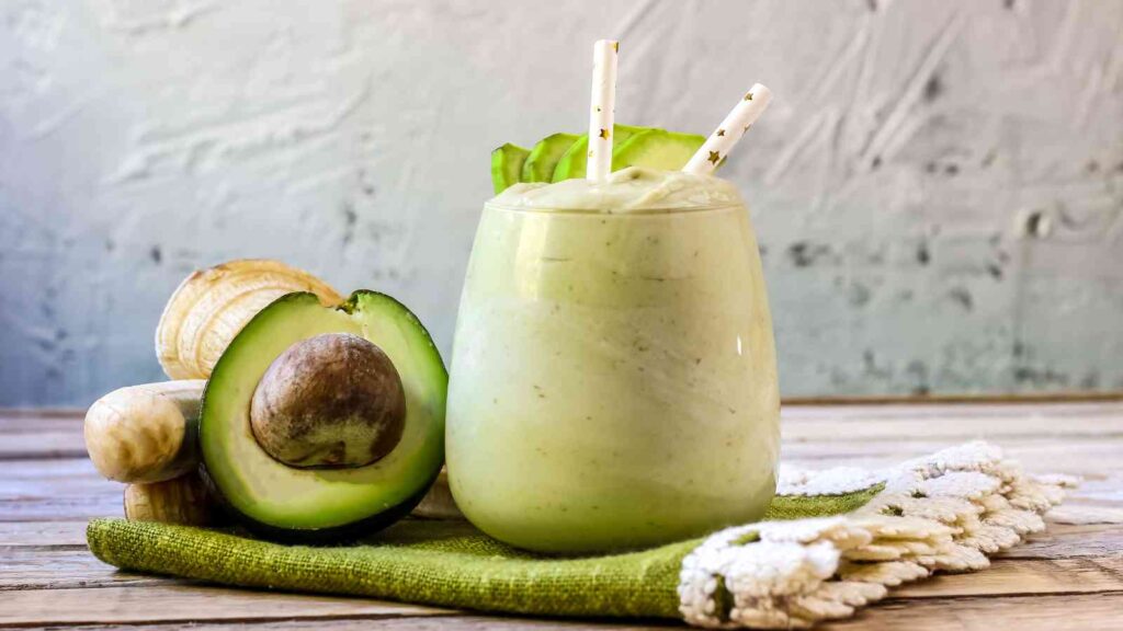 A glass of creamy green smoothie with two straws sits beside a halved avocado and a bunch of bananas on a green cloth.