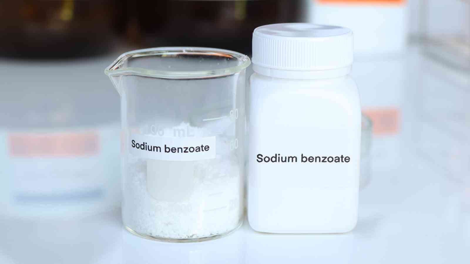 A beaker and a plastic bottle, both labeled "Sodium benzoate," contain a white powder, set on a laboratory surface.