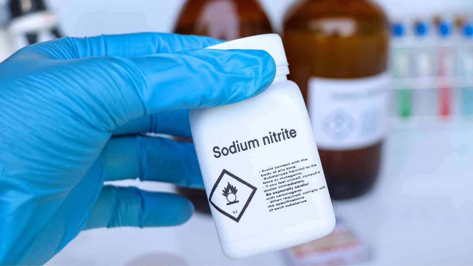 A gloved hand holds a white plastic bottle labeled "Sodium nitrite" with a hazard symbol, with laboratory bottles and equipment blurred in the background.