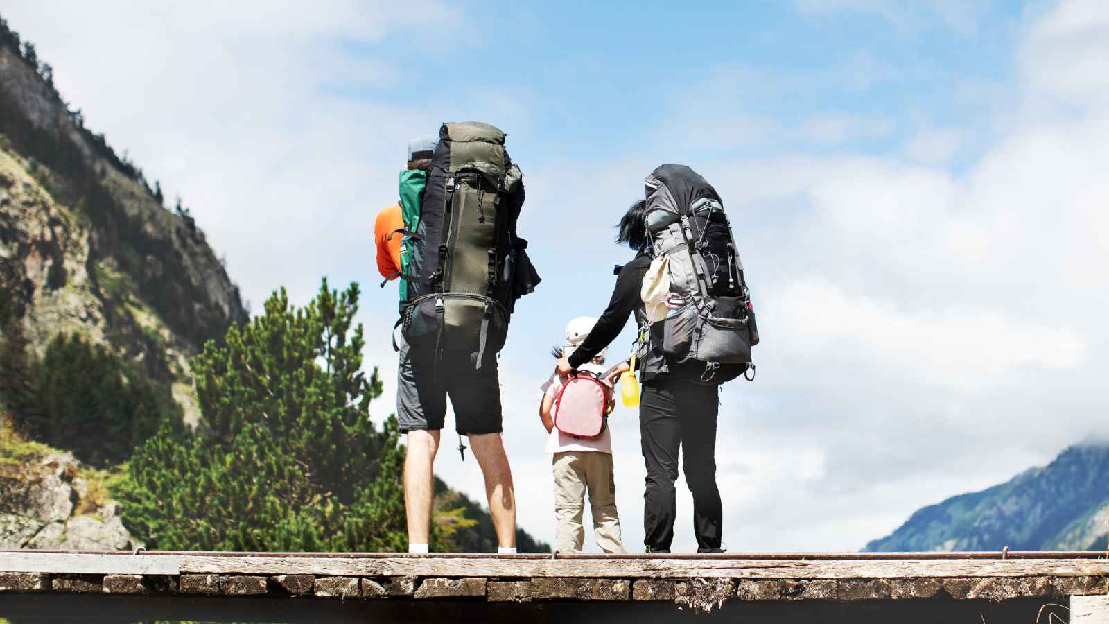 Three people with large backpacks stand on a wooden platform outdoors, looking toward mountains and trees under a partly cloudy sky.