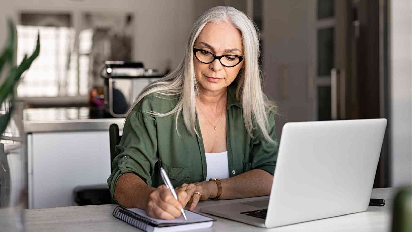 Woman with long gray hair and glasses writes in a notebook while working on a laptop at a desk in a modern home setting.