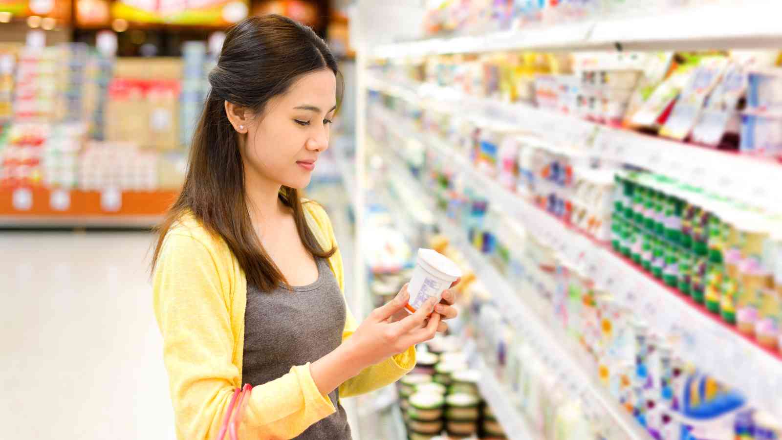 A woman stands in a grocery store aisle, reading the label on a product from the refrigerated section, with shelves of various dairy items in the background.