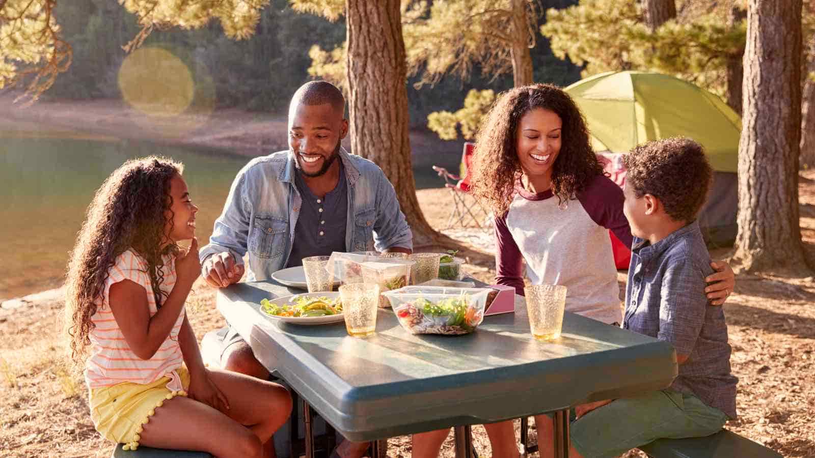 A family of four sits at an outdoor picnic table, smiling and eating a meal together near a tent and trees at a campsite.