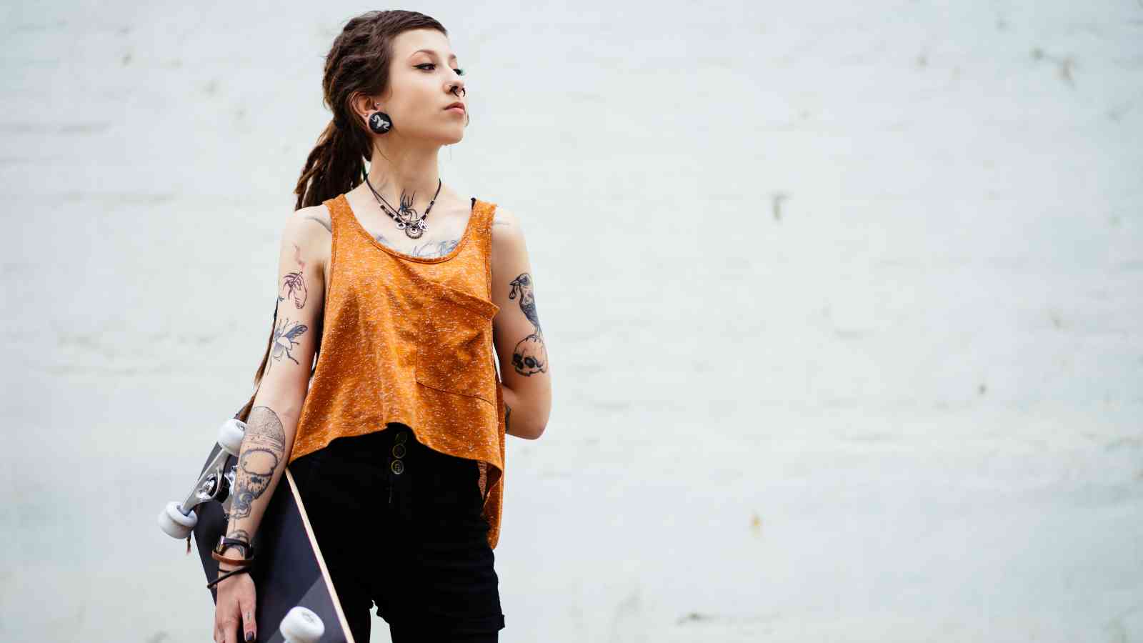 Person with tattoos and piercings, wearing an orange sleeveless top and black shorts, holds a skateboard while standing against a plain light-colored wall.