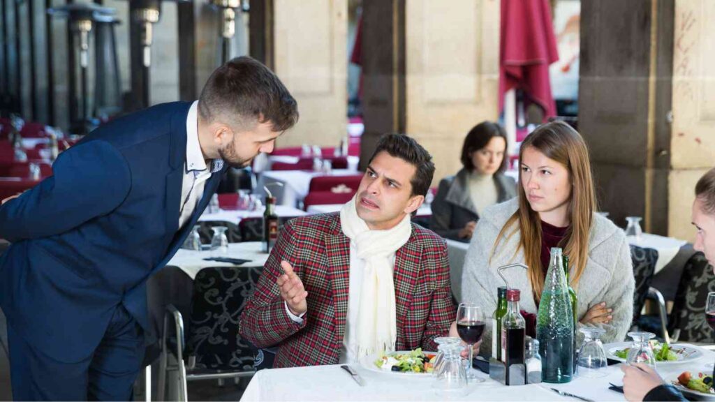 A man in a plaid jacket gestures while talking to a waiter at an outdoor restaurant. A woman sits beside him, looking on. There are drinks and plates of food on the table.