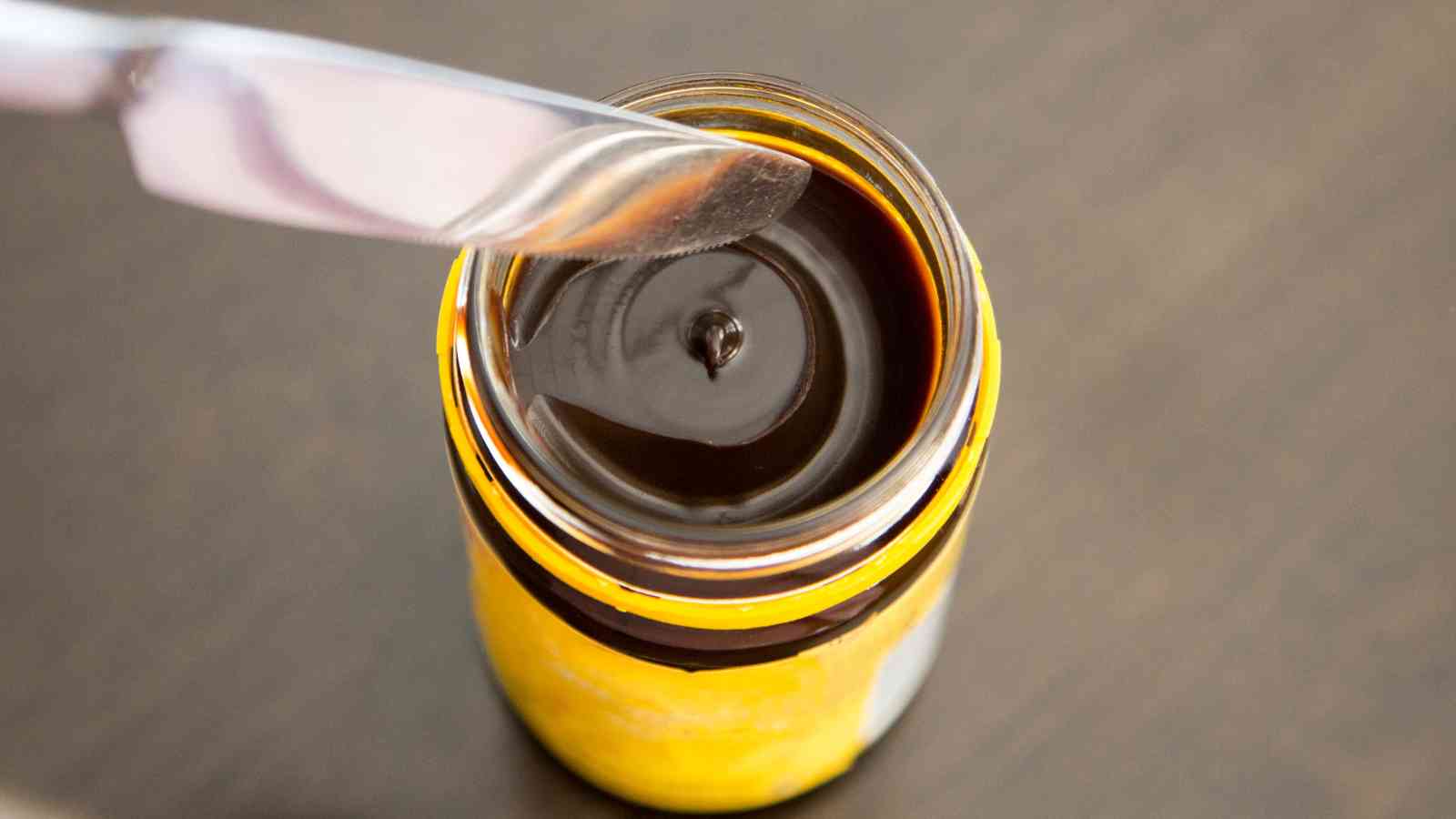 A knife hovers over an open jar filled with dark brown spread, viewed from above, on a dark surface.
