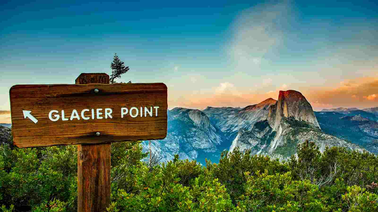 Wooden sign marked "Glacier Point" with an arrow, set against a scenic mountain landscape at sunset, including a prominent granite peak.