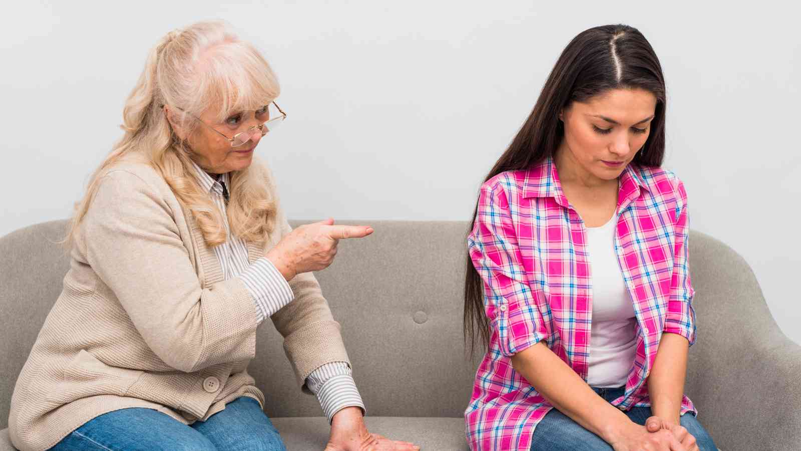 Older woman pointing and talking to a younger woman who looks upset and is sitting with her head bowed on a sofa.