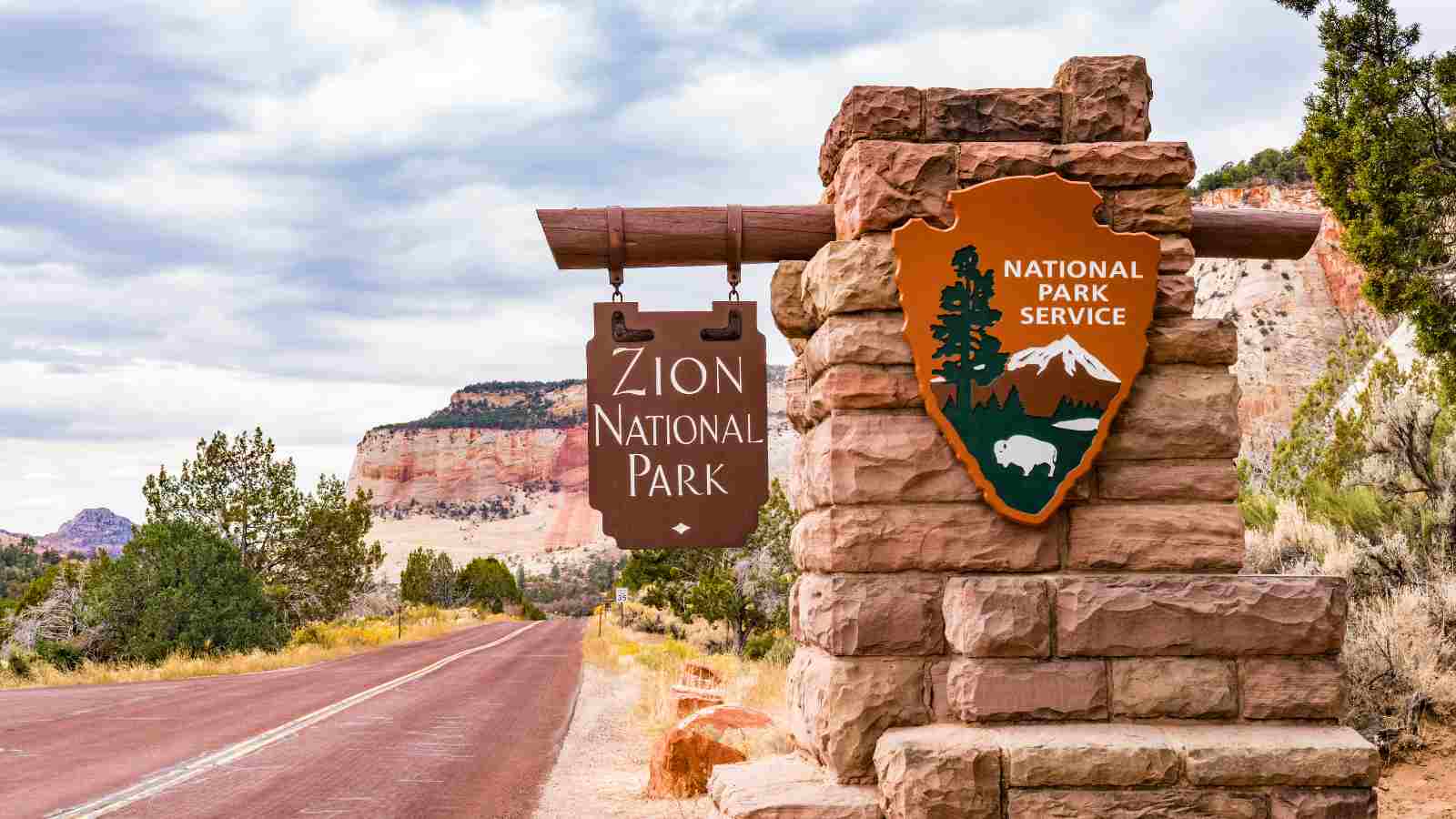 Stone entrance sign for Zion National Park beside a road, with trees and rocky hills in the background under a cloudy sky.