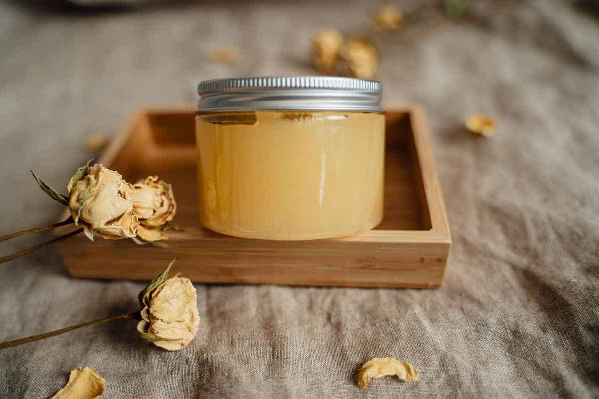 A glass jar with a silver lid filled with a yellow substance sits in a wooden tray, surrounded by dried yellow roses on a beige fabric surface.