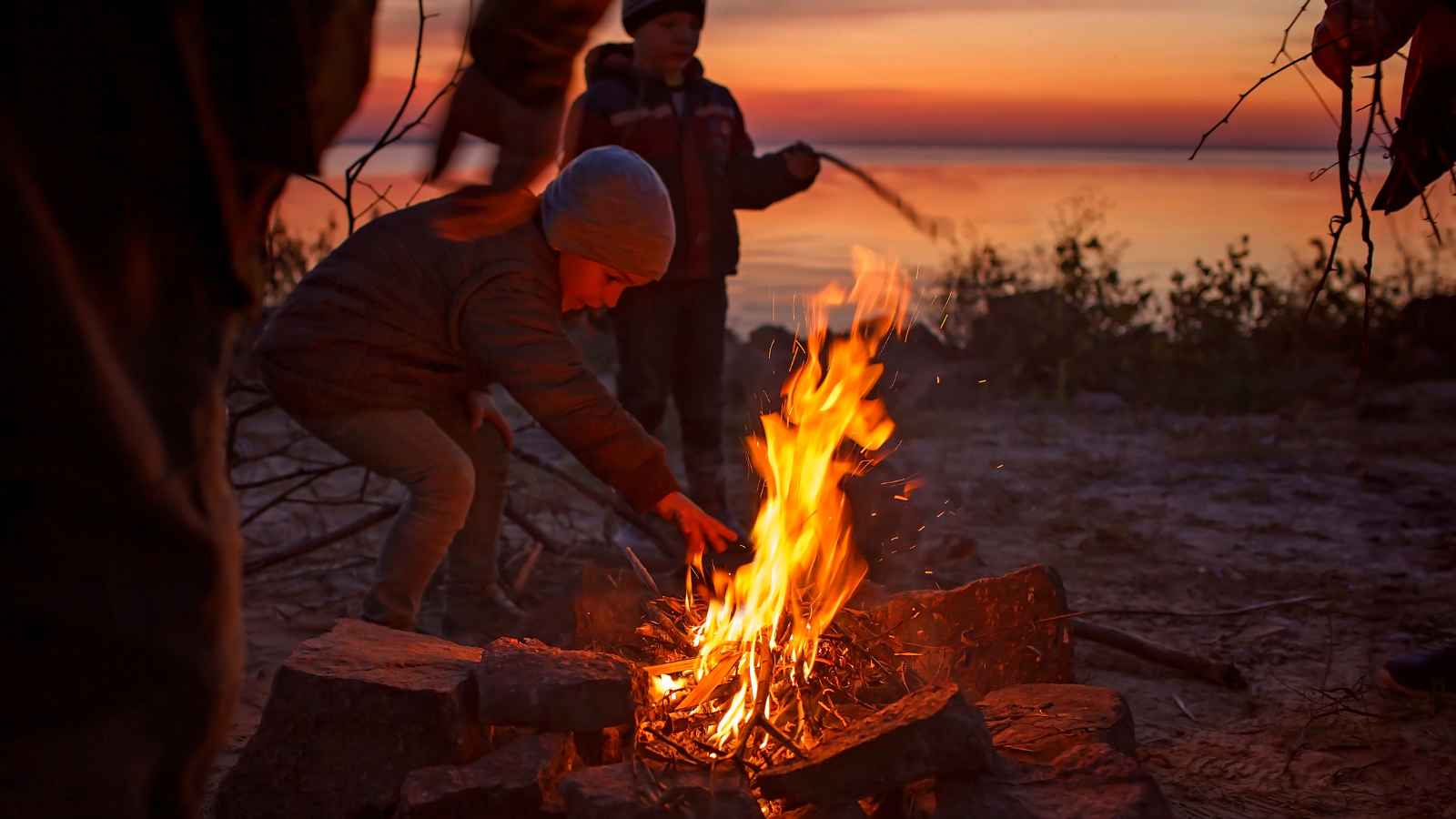 A child wearing a hat tends to a campfire on a beach at sunset, with other children and a body of water in the background.