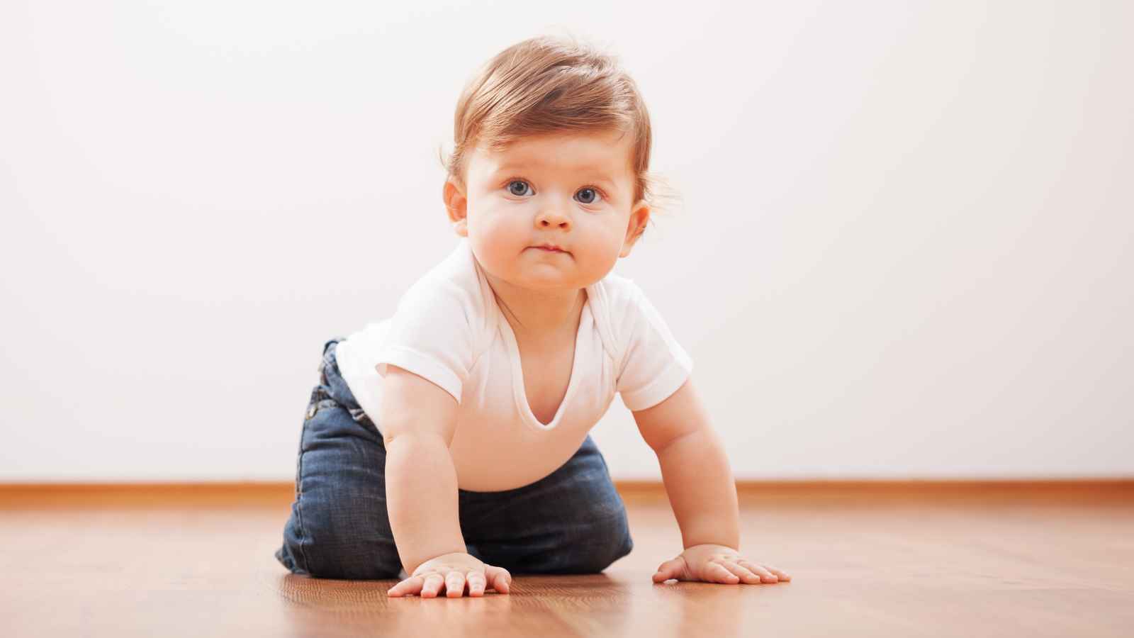 A baby with light brown hair wearing a white shirt and jeans is crawling on a wooden floor, looking forward.