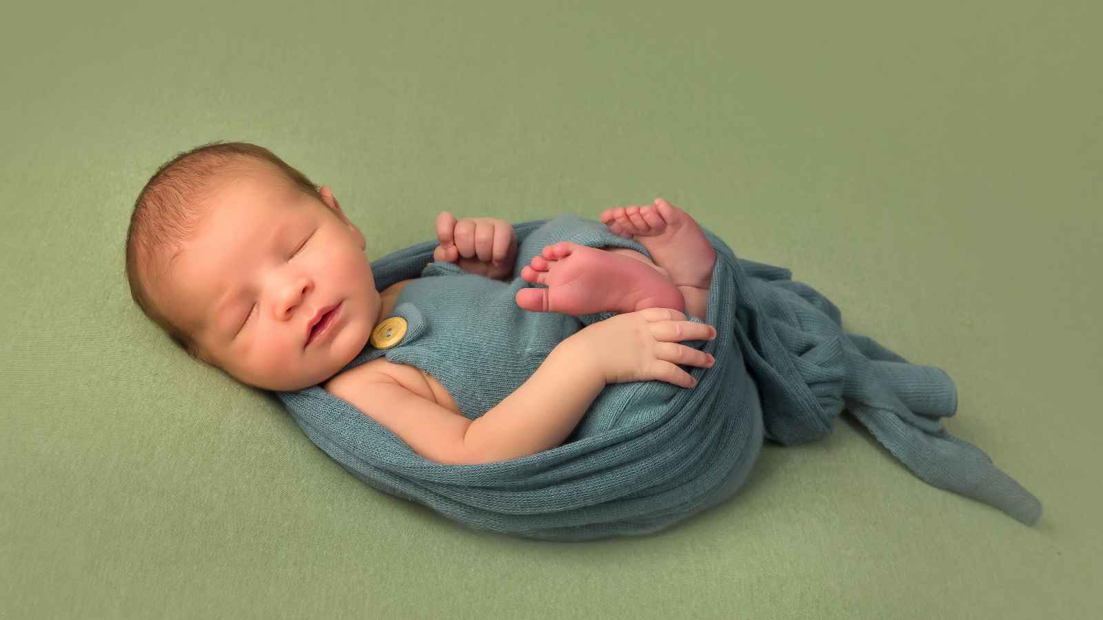 A newborn baby wrapped in a blue swaddle sleeps on a light green surface, with hands and feet visible.