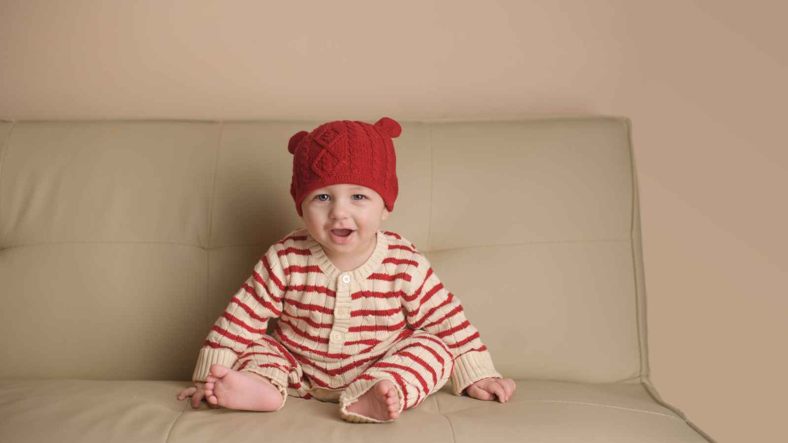 A baby wears a red knit hat and red-and-white striped pajamas while sitting barefoot on a beige sofa against a plain wall.