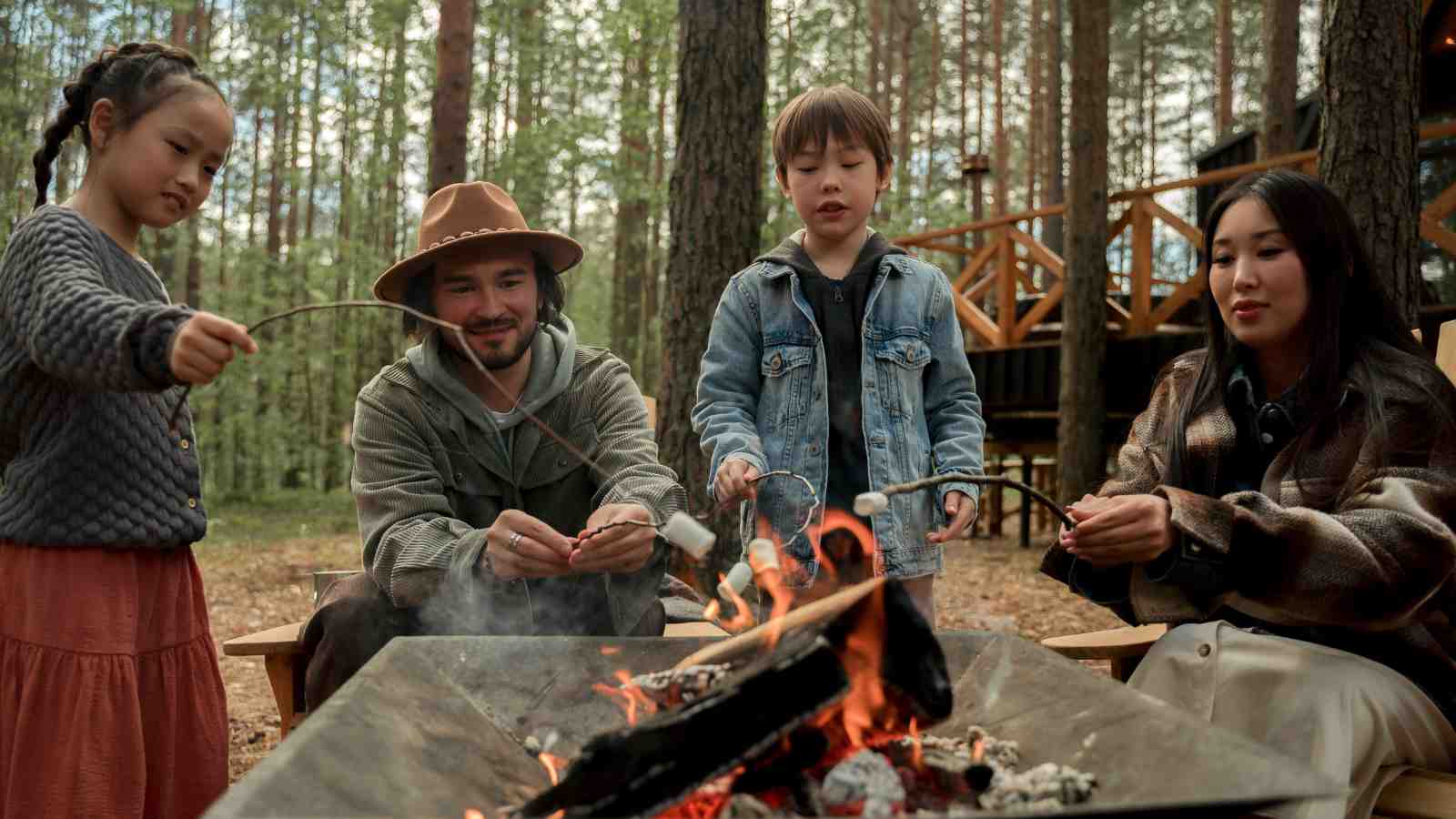 Four people sit around a campfire in a forest, roasting marshmallows on sticks. Wooden cabins are visible in the background among the trees.
