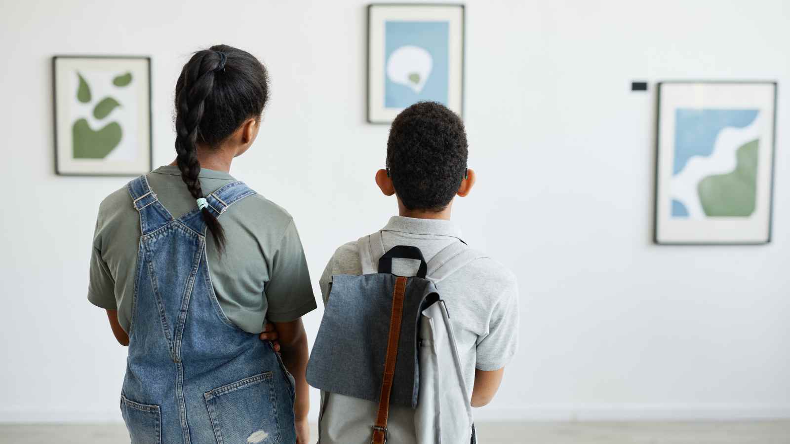 Two children with backpacks stand side by side, looking at framed abstract art on a white gallery wall.