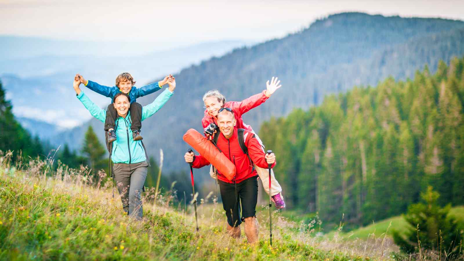Four people, two adults and two children, hike up a grassy hill with forested mountains in the background. One child rides on each adult’s back. All appear to be smiling.