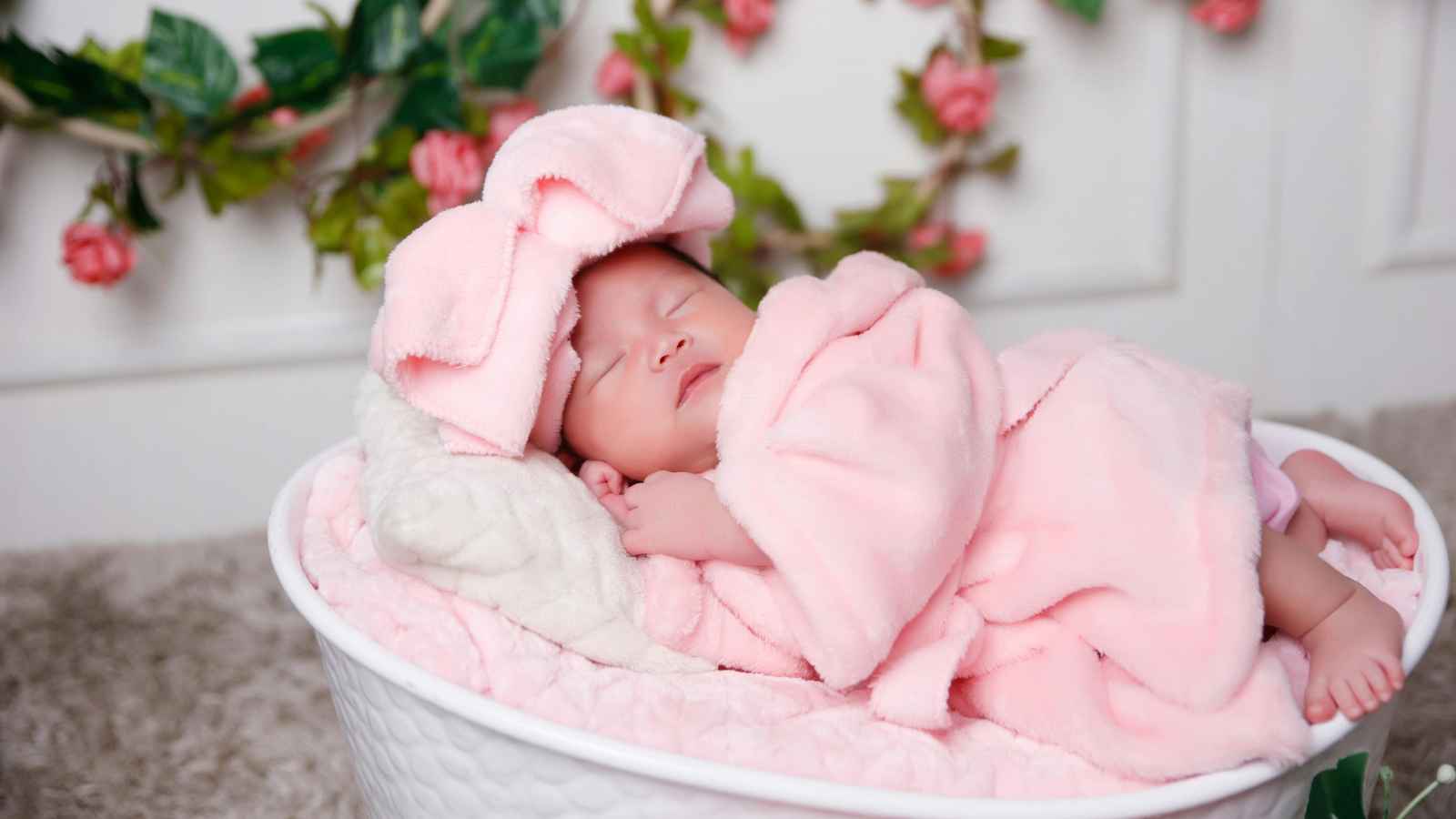 A newborn baby sleeps in a white basket, wrapped in a pink blanket with a large pink bow on her head; as pink flowers and greenery decorate the background.