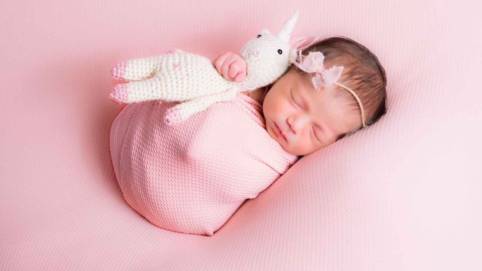 A newborn baby wrapped in a pink blanket sleeps on a pink surface, holding a small knitted unicorn toy and wearing a headband with a flower.