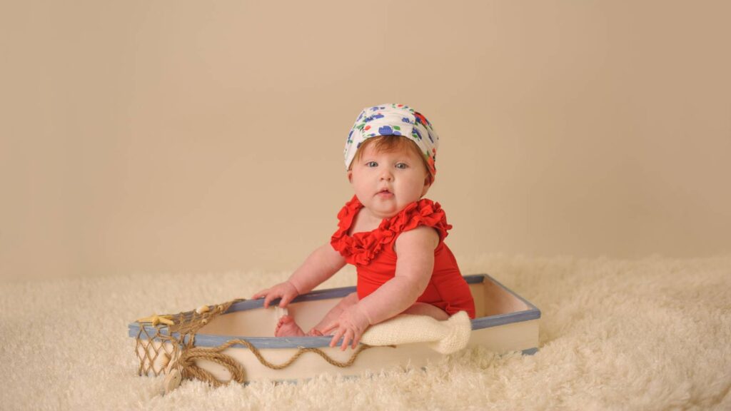 A baby wearing a red outfit and patterned hat sits in a small wooden boat prop on a soft beige surface, looking toward the camera.