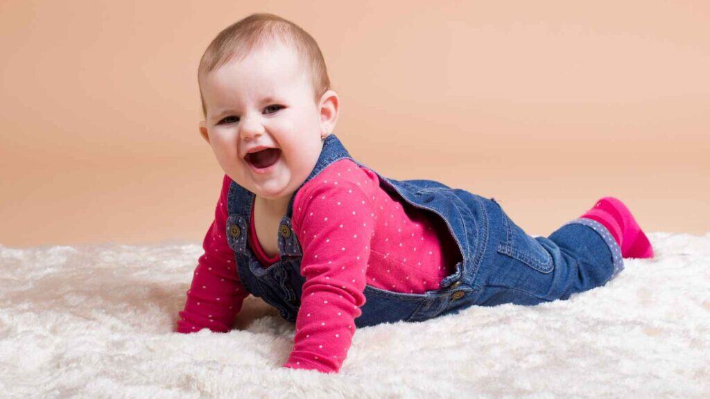 A baby girl wears a pink shirt with white polka dots and denim overalls as she lies on a soft white rug, smiling with her mouth open.
