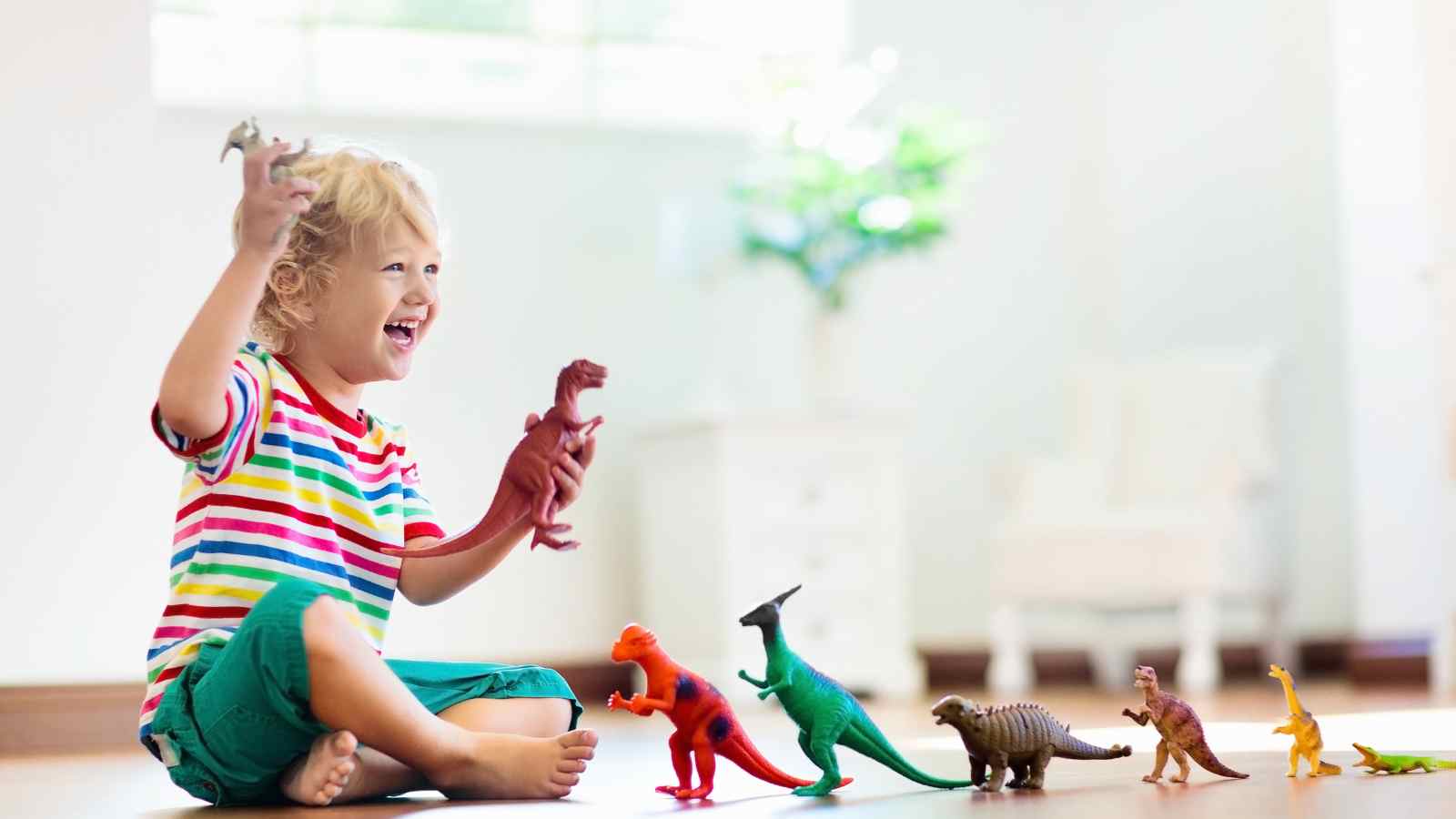 A young child in a striped shirt sits on the floor, smiling and playing with various colorful toy dinosaurs arranged in front of them.