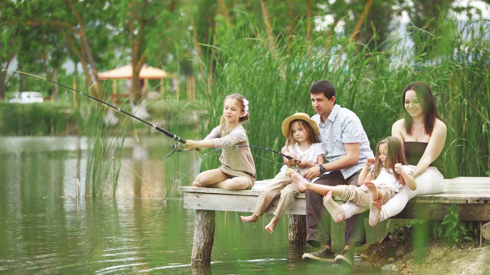 A family of four sits on a wooden dock by a lake, with one child fishing while the others sit nearby, surrounded by green trees and tall grass.