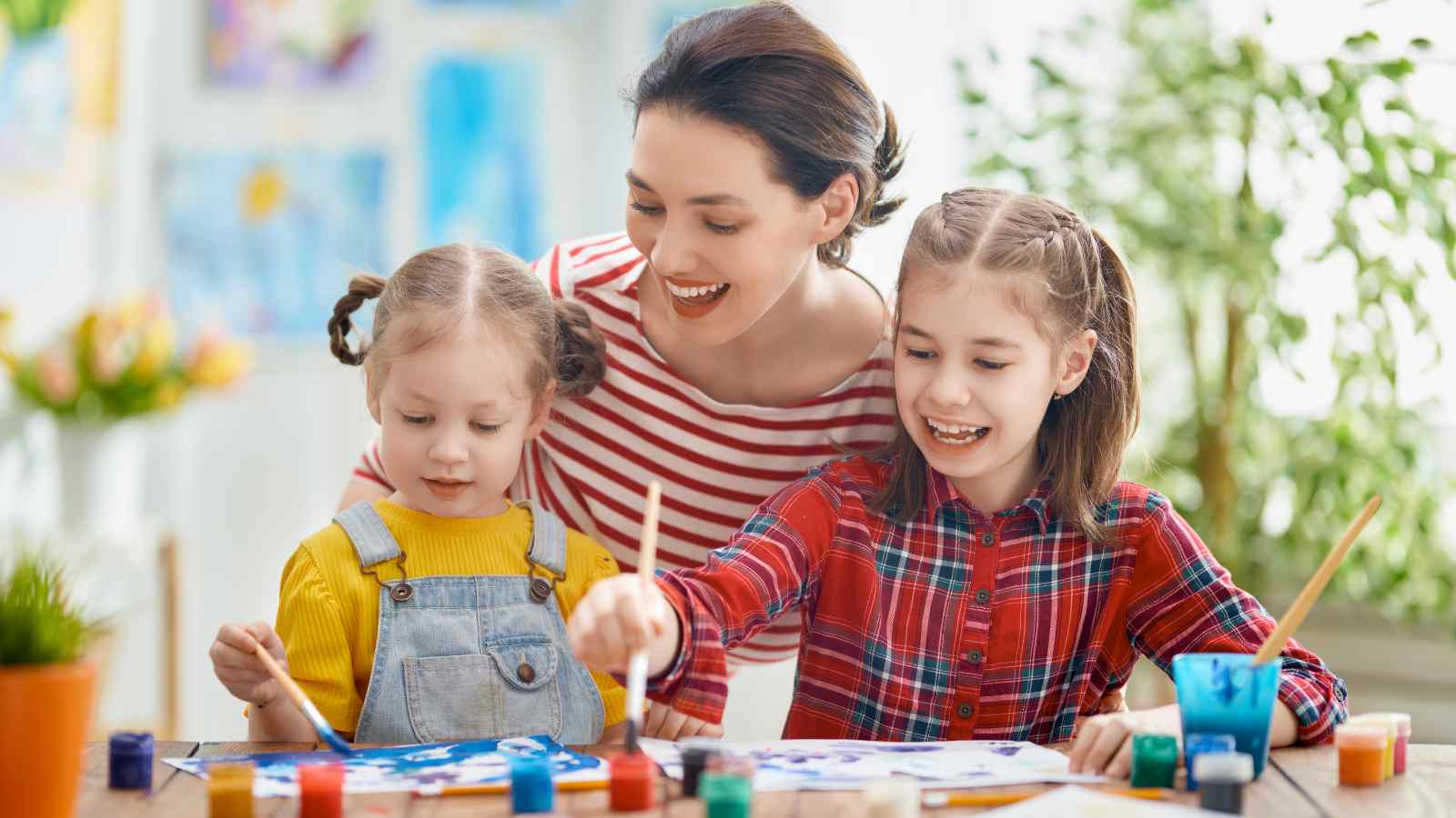 An adult and two young girls sit at a table painting with brushes and colorful paints, smiling and engaged in an art activity.