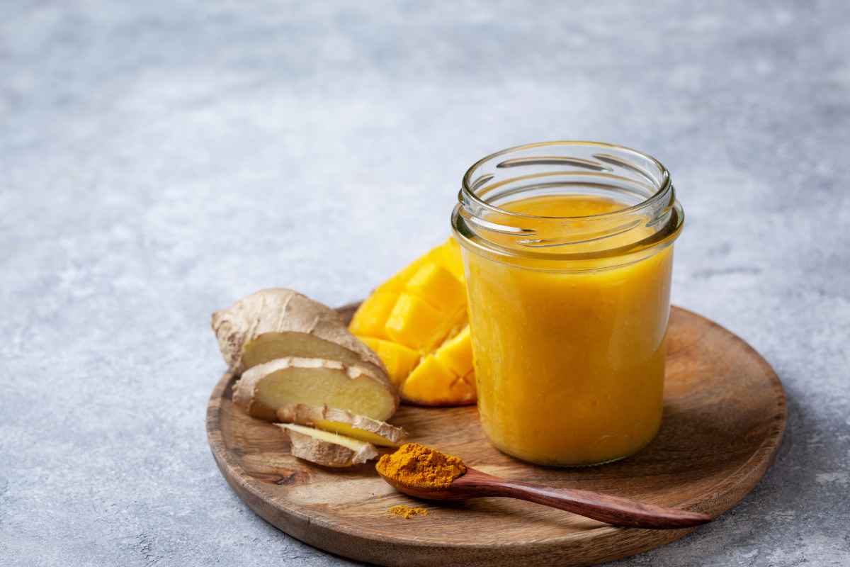 A jar of yellow smoothie sits on a wooden plate with sliced ginger, mango pieces, and a wooden spoon holding vibrant turmeric powder.