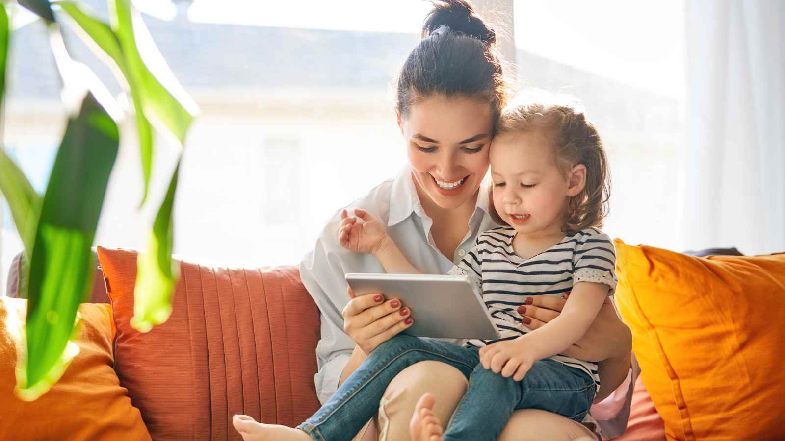 A woman and a young girl sit on a couch, smiling and looking at a tablet together in a brightly lit room.