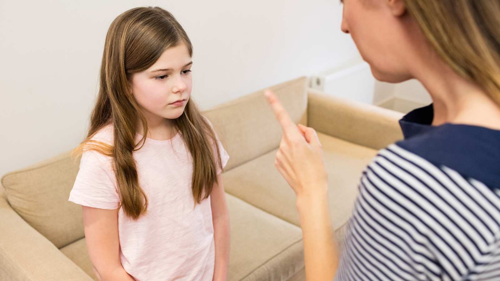 An adult is pointing a finger while talking to a young girl who looks downcast, standing in front of a beige couch indoors.